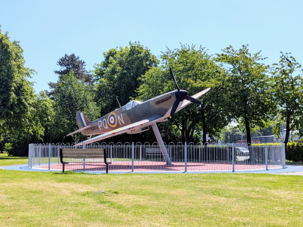 Spitfire replica against a bank of trees, surrounded by a metal fence.